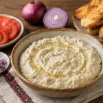 Salata de vinete cremoasa intr-un bol, servita cu rosii, ceapa rosie si paine prajita.